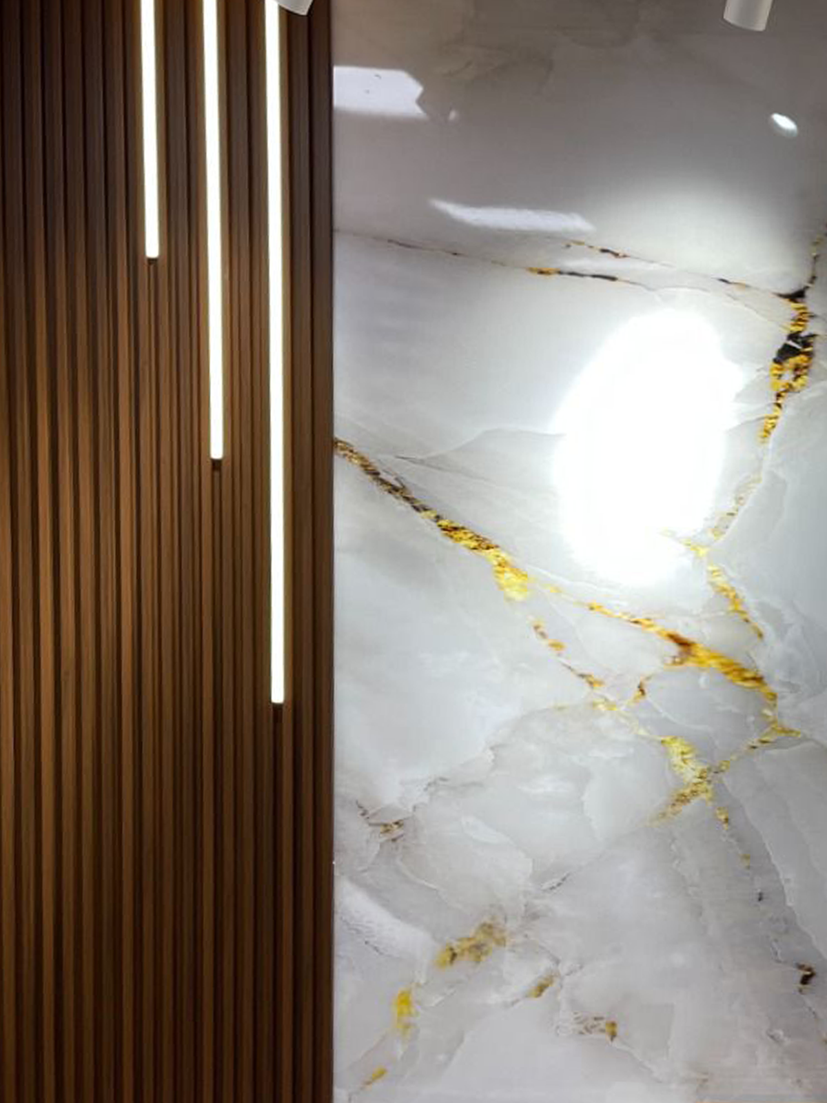 Vertical LED strip accent lighting installed on wood fluted wall panels beside luxury white marble wall surface with gold veining, showcasing modern interior wall lighting design